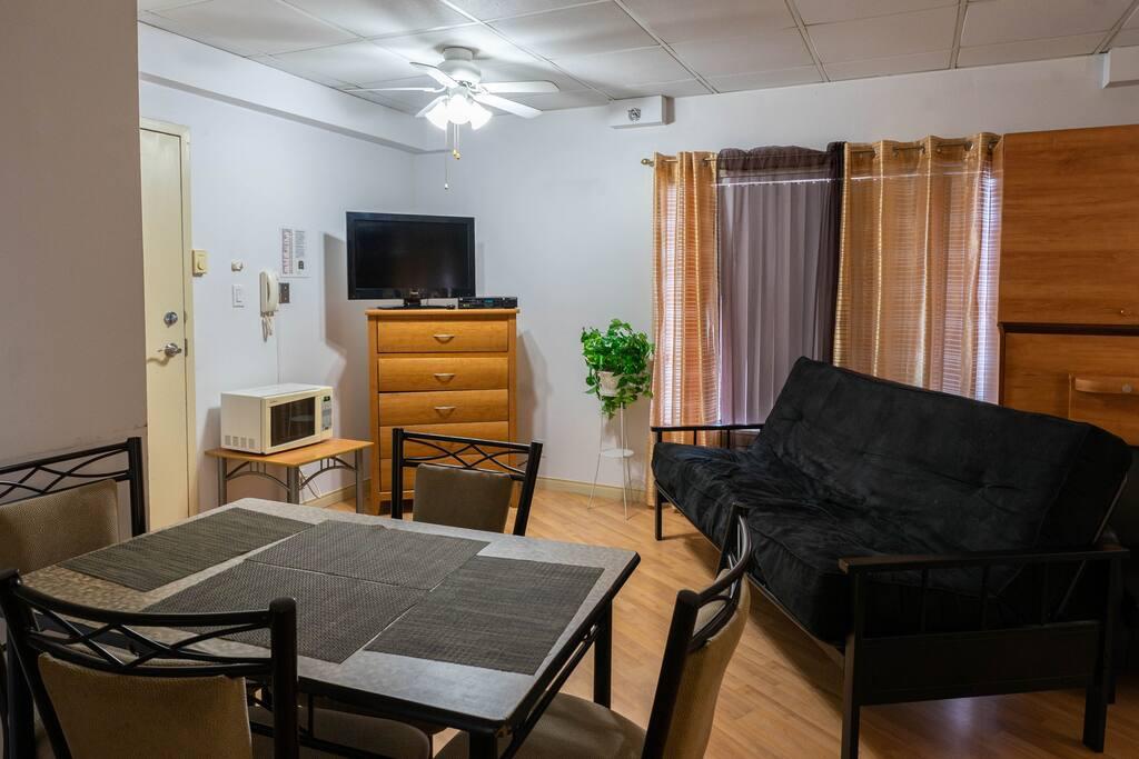 Living area with a black futon, a dresser with a TV, a potted plant and a dining table for four