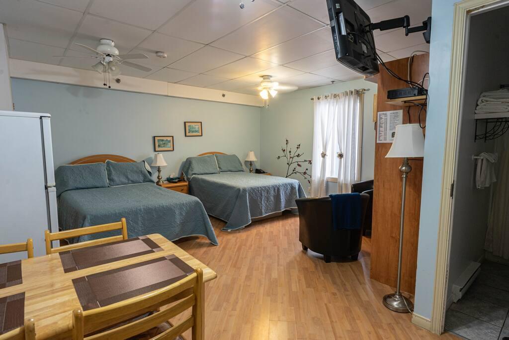 Sage-green room with two beds, a round wood table and a kitchenette glimpsed through an open doorway