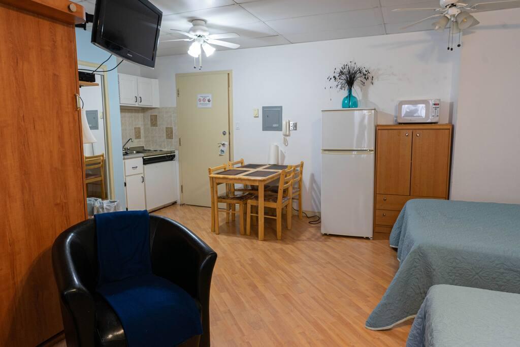Room with a wood wardrobe, a kitchenette glimpsed through a doorway, a dining table and beds with sage-green bedspreads
