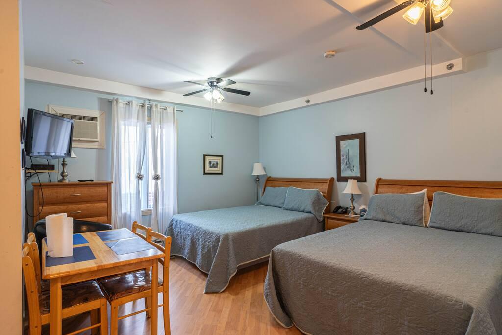 Pale blue room with two beds with grey-blue bedspreads, a window air conditioner and a small dining table