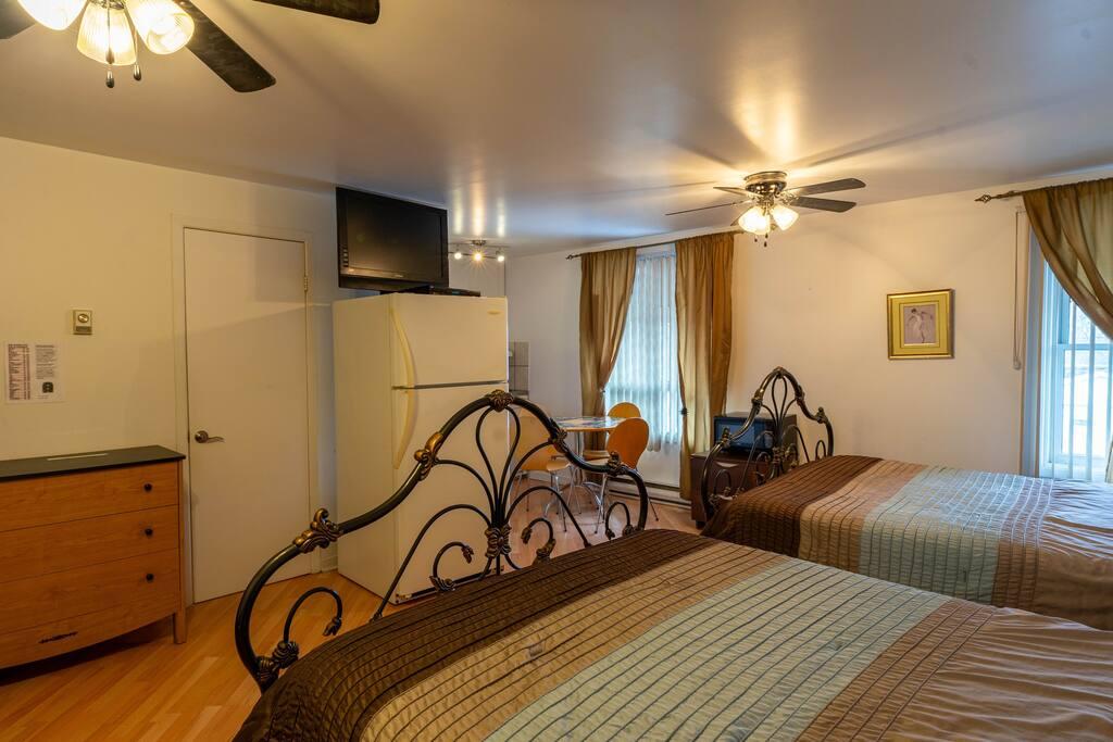 Room with two wrought-iron bed frames, brown and green striped bedspreads, and a refrigerator topped with a TV