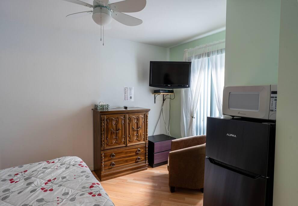 Room with an ornate wood chest, a wall-mounted TV, a mini-fridge with a microwave and a floral-patterned bed