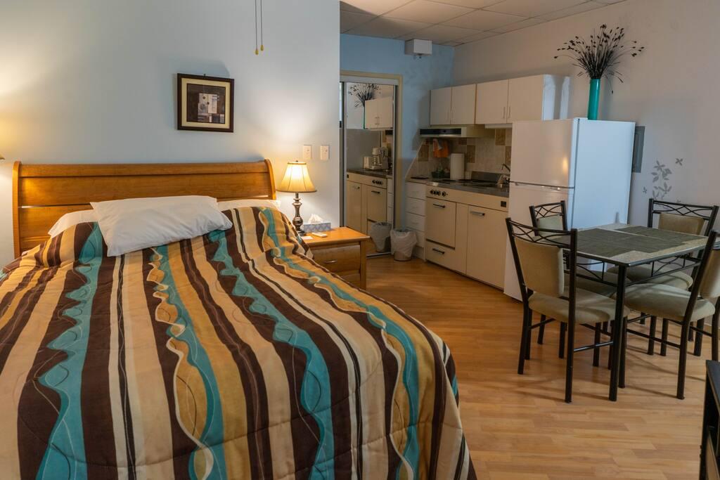 Room with a bed with a brown and teal wave-patterned bedspread, a kitchenette and dining table visible in the background
