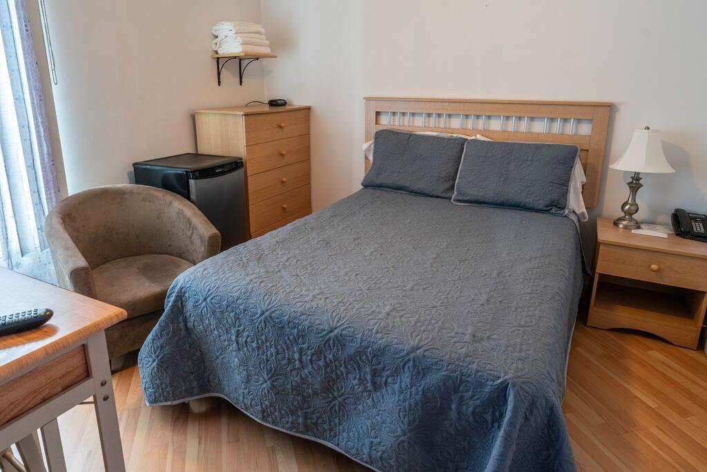 Room with a slatted wood headboard bed, a blue-grey bedspread, a mini-fridge and a suede armchair