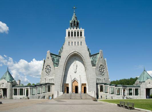 Façade en pierre grise du Sanctuaire Notre-Dame-du-Cap sous un ciel bleu, avec son clocher central et ses deux ailes à toit vert-de-gris
