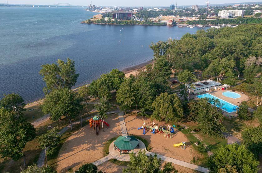 Vue aérienne du parc de l'île Saint-Quentin au bord du fleuve, avec ses jeux pour enfants, son kiosque à toit vert et sa piscine extérieure