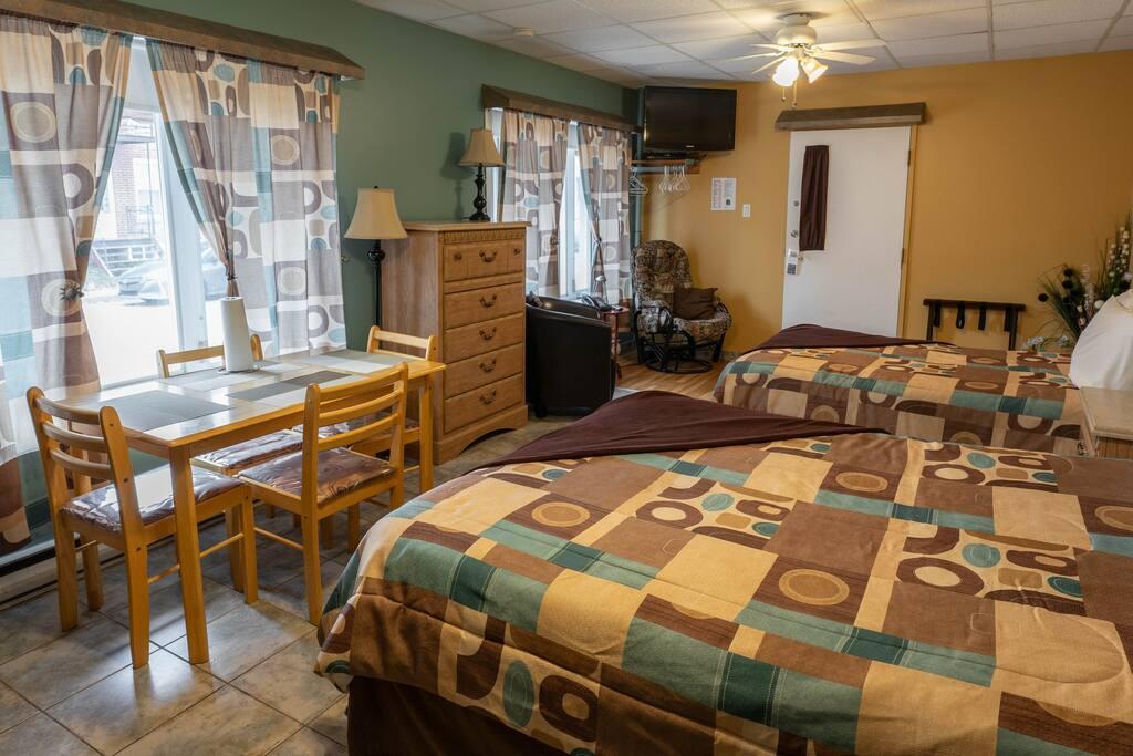 Chambre avec deux lits doubles à couvre-lits à motifs, table à manger pour quatre près de la fenêtre et commode en bois