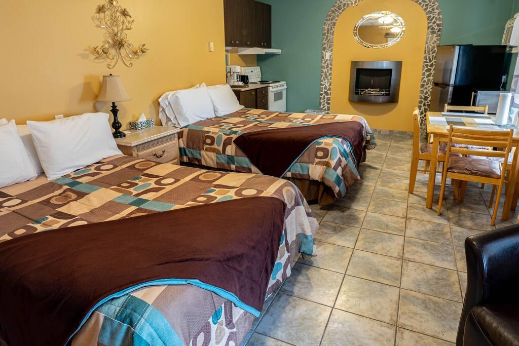 Chambre avec deux lits, foyer électrique dans une alcôve décorative, cuisinette et table à manger visibles en arrière-plan