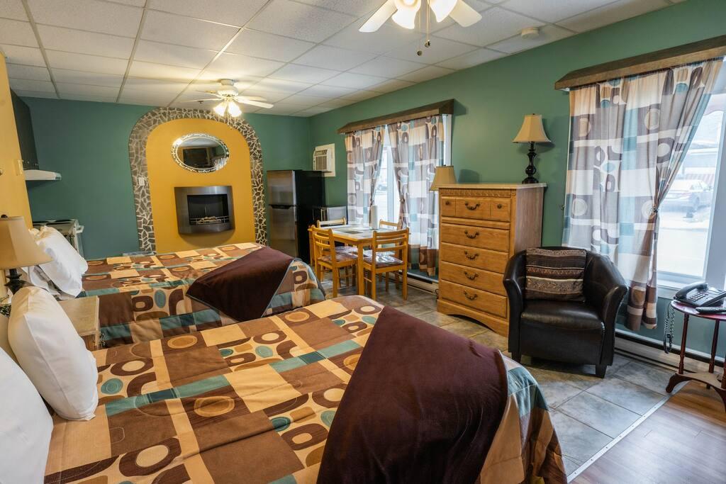 Chambre avec deux lits, foyer électrique sous arche en pierre décorative, cuisinette avec réfrigérateur et espace repas