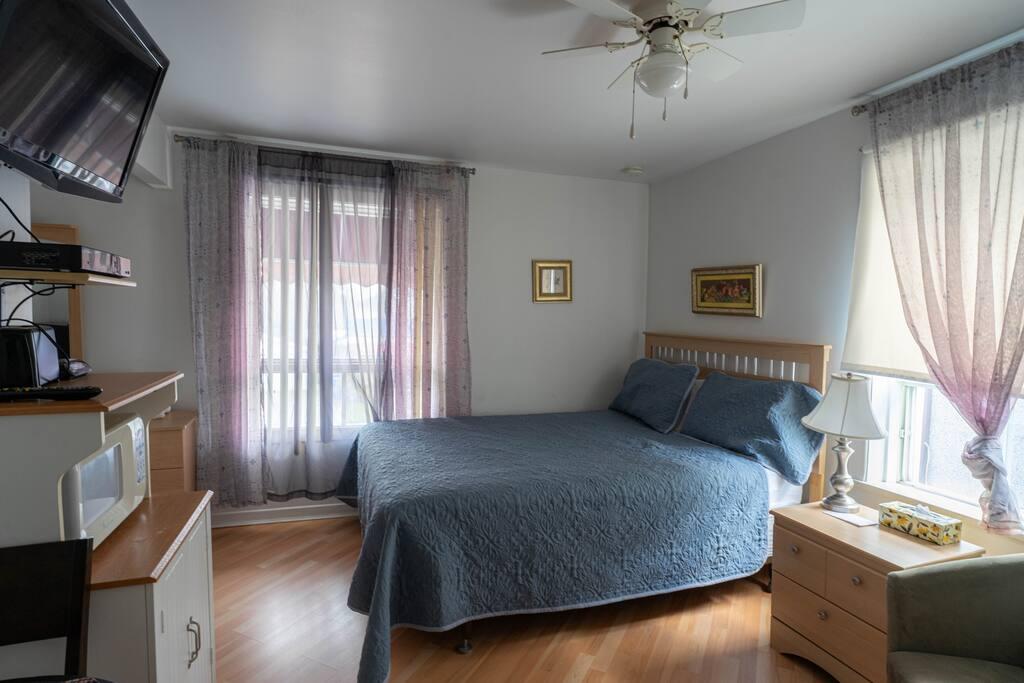 Chambre avec lit à couvre-lit bleu, rideaux mauves translucides et meuble multimédia avec four à micro-ondes