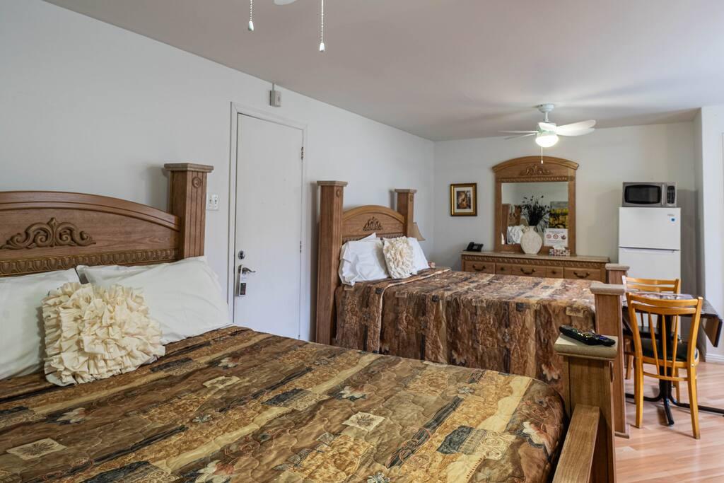Chambre avec deux lits en bois sculpté, couvre-lits à motifs bruns, commode-miroir et coin repas avec réfrigérateur