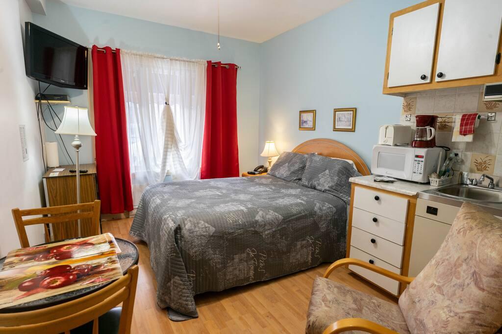 Chambre bleu pâle avec rideaux rouges, cuisinette et lit à couvre-lit gris, commode avec téléviseur